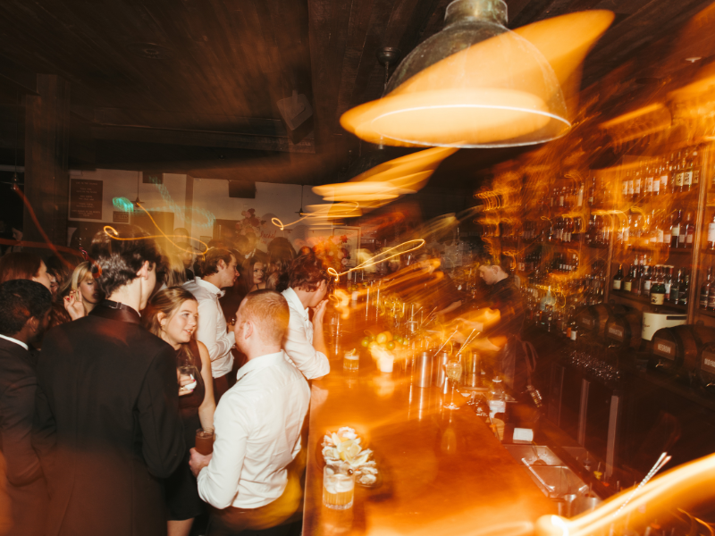 People gathered at a bar or party, with warm lighting and a lively atmosphere, featuring a long bar counter and bottles on shelves.