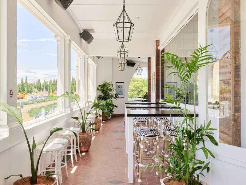 Highball bright enclosed terrace area with large windows, potted plants, wooden bar stools, and hanging lantern-style lights