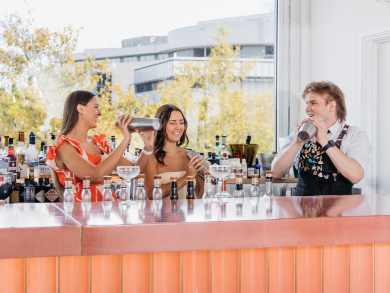 Three people behind a bar smiling and drinking cocktails, with bottles of alcohol on the bar and a large window showing trees and buildings outside.