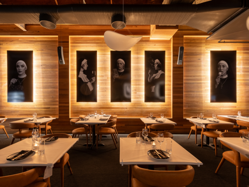 Interior of a modern restaurant with wooden walls, illuminated black-and-white portrait photographs of women, and neatly set tables with plates, glasses, and silverware.