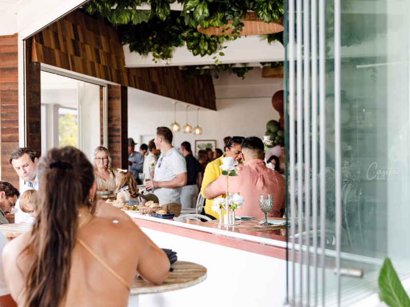People gathered in a modern, well-lit indoor space with a view from outside, socializing and dining, with plants and wooden accents.