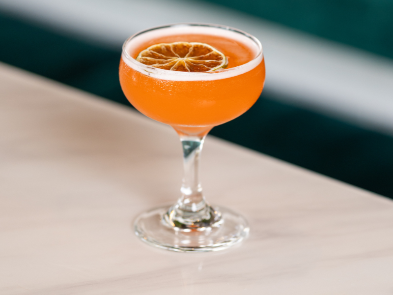 A cocktail glass with an orange-colored drink and a slice of dried citrus fruit garnish.