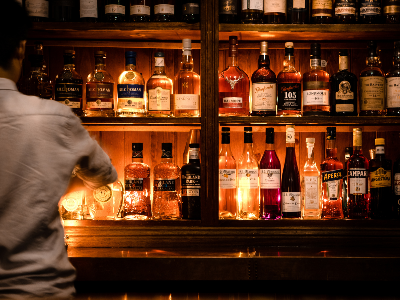Bar with shelves of various bottles of liquor, illuminated with warm lighting, and a person in a light-colored shirt standing in front.