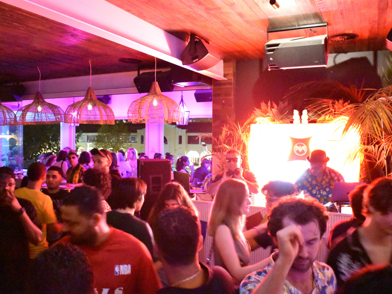 Crowded nightclub with people dancing and socializing, illuminated by colorful purple and pink lighting, with a DJ booth and tropical plants in the background.