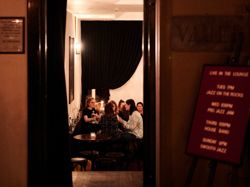 A group of young women sitting at a table inside a dimly lit lounge or bar, engaged in conversation, with glasses and drinks on the table.