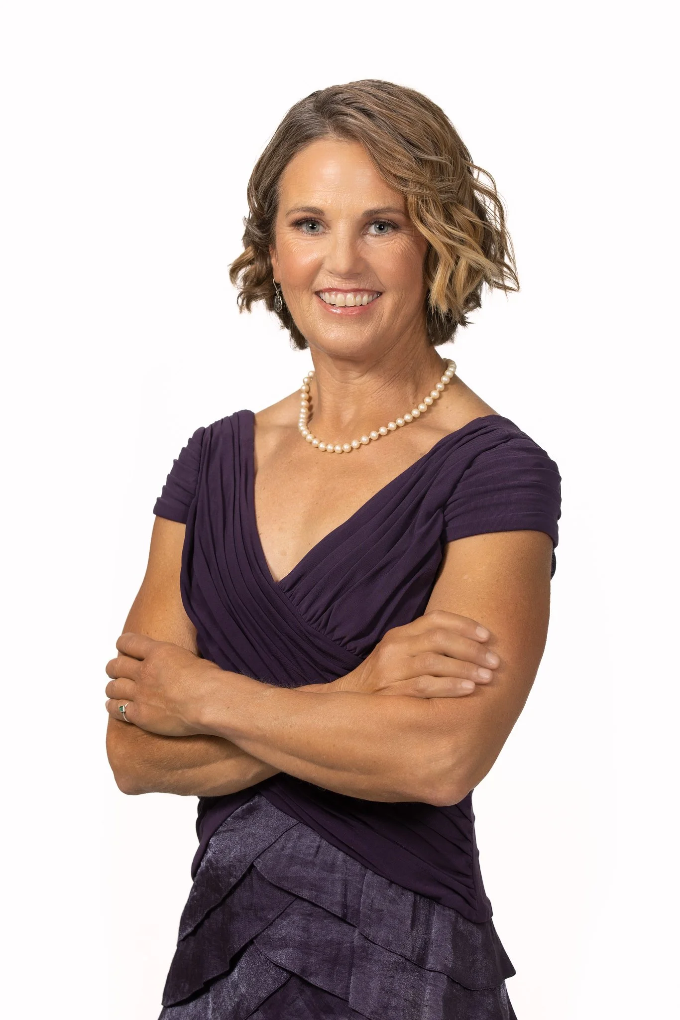 Portrait of a woman with short wavy brown hair, wearing a purple dress, pearl necklace, and arms crossed, smiling against a white background.