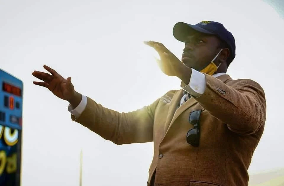 A man wearing a brown jacket, a baseball cap, and sunglasses hanging from his shirt is gesturing with his hands outdoors against a bright sky.