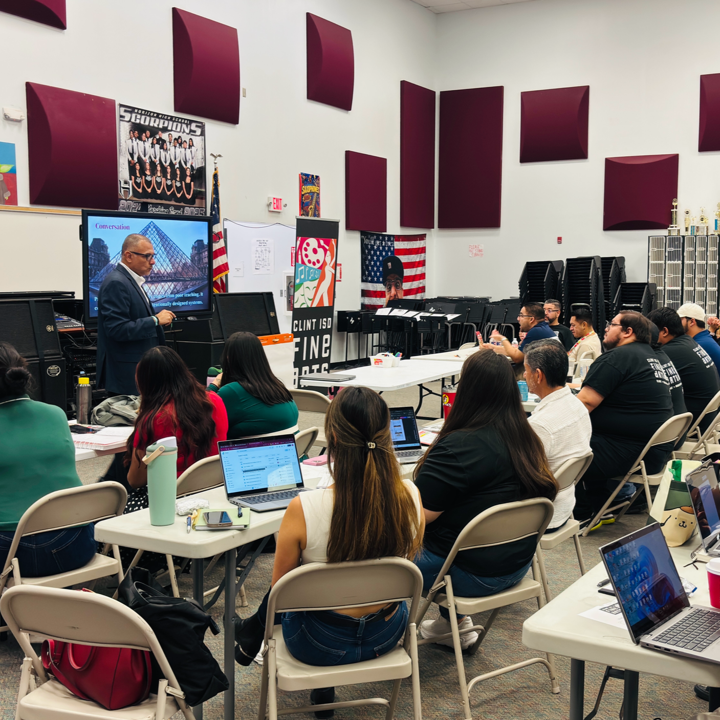 A classroom or conference room with a group of people seated at tables, listening to a male presenter in a suit. The room has white walls with maroon acoustic panels and various posters, including a large one of a school group and an American flag. Attendees have laptops, notebooks, and drinks. The presenter stands near a screen displaying a presentation slide.