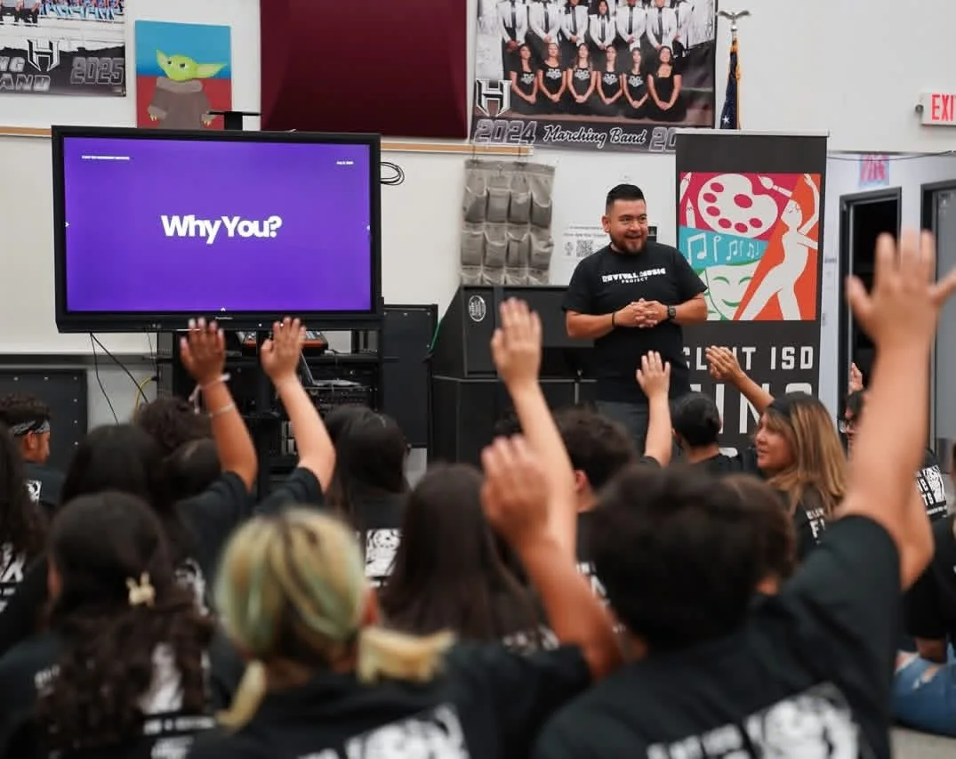 A classroom or assembly hall with students raising hands while a man stands at the front near a TV screen displaying 'Why You?'. The classroom has posters, banners, and a backdrop with 'Marching Band 2024' on the wall.