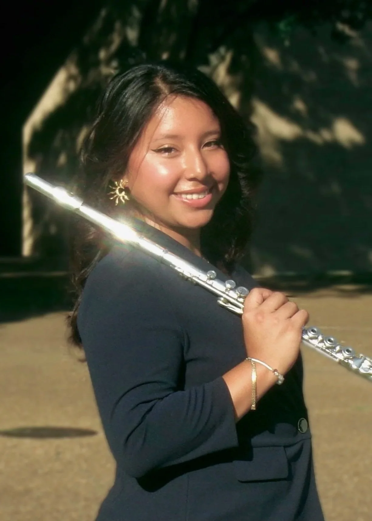 A young woman with dark hair holding a silver flute on her shoulder, smiling at the camera, outdoors with trees in the background.
