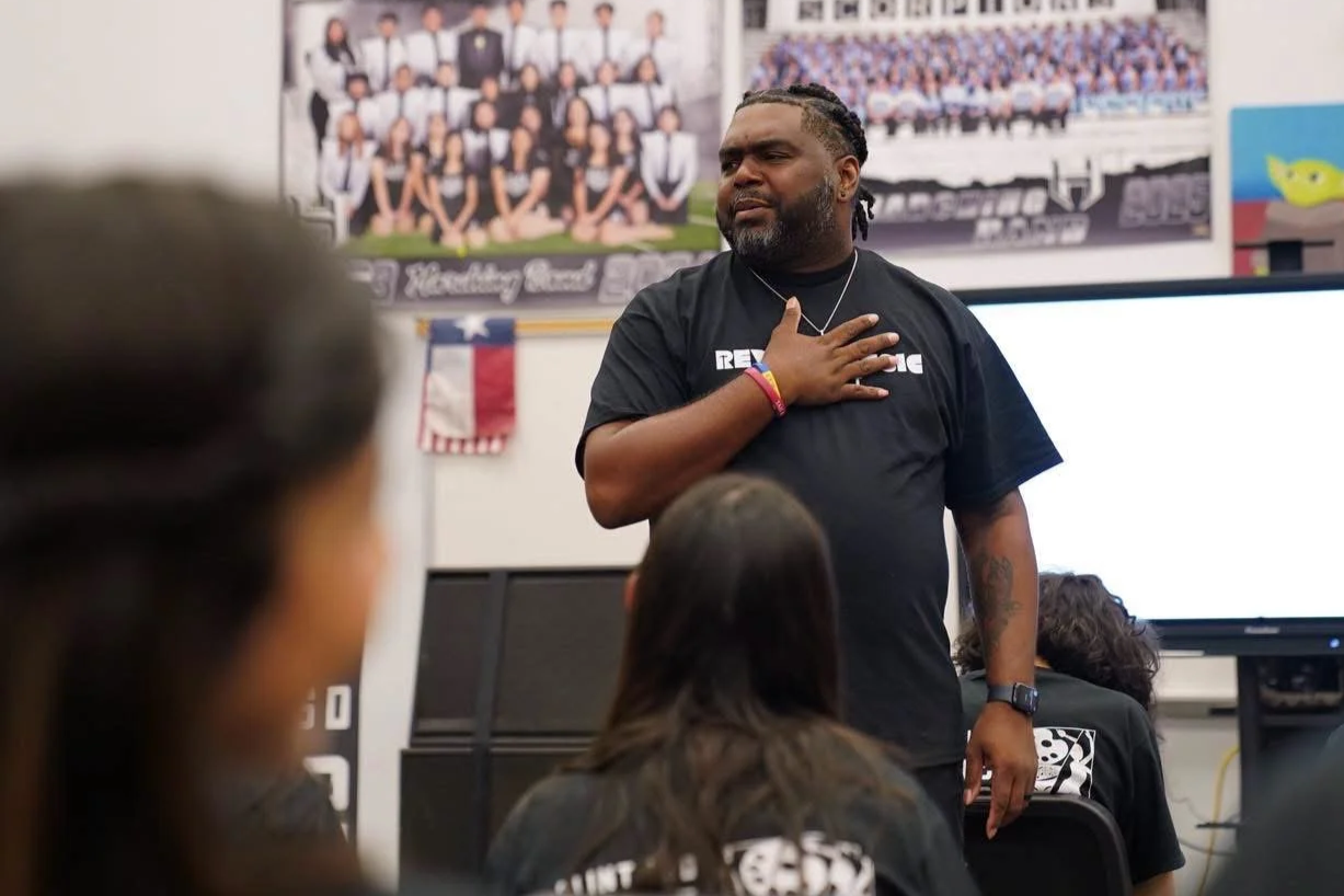 A man with a beard and braided hair stands in a classroom, placing his right hand on his chest, speaking to students. Students are facing him. There are posters and a Texas flag on the wall behind him.