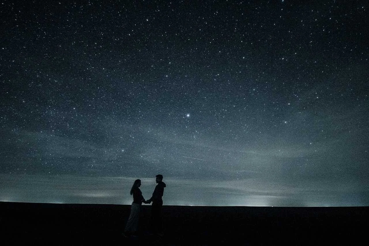 Un par de personas sosteniéndose de las manos en un campo, con un cielo estrellado y la Vía Láctea en el fondo.