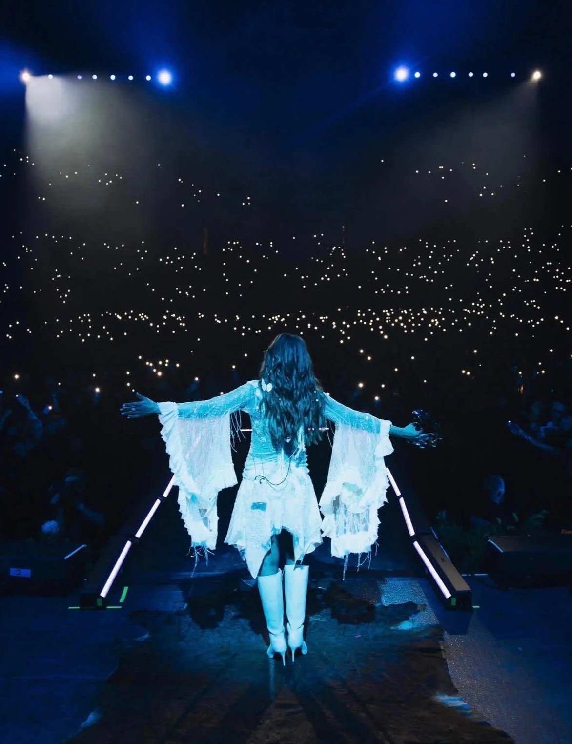 A performer standing on stage with arms outstretched facing a large audience in a dark concert hall, illuminated by blue and white stage lights, with audience members holding up cell phones.