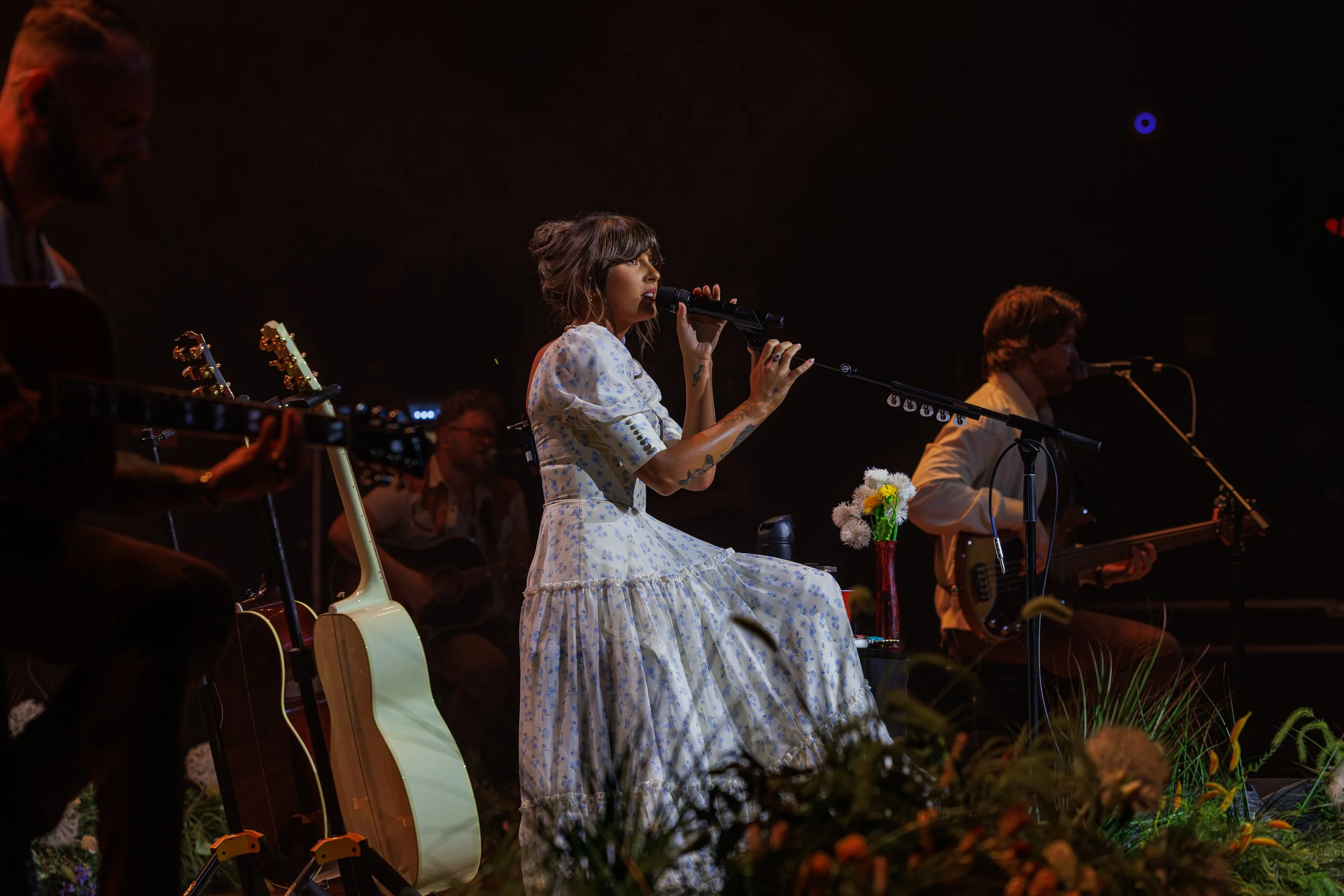 A woman in a white floral dress is singing into a microphone on stage. She is surrounded by musicians playing guitars and other instruments, with stage lighting creating a warm, intimate atmosphere. Flowers are visible in the foreground.