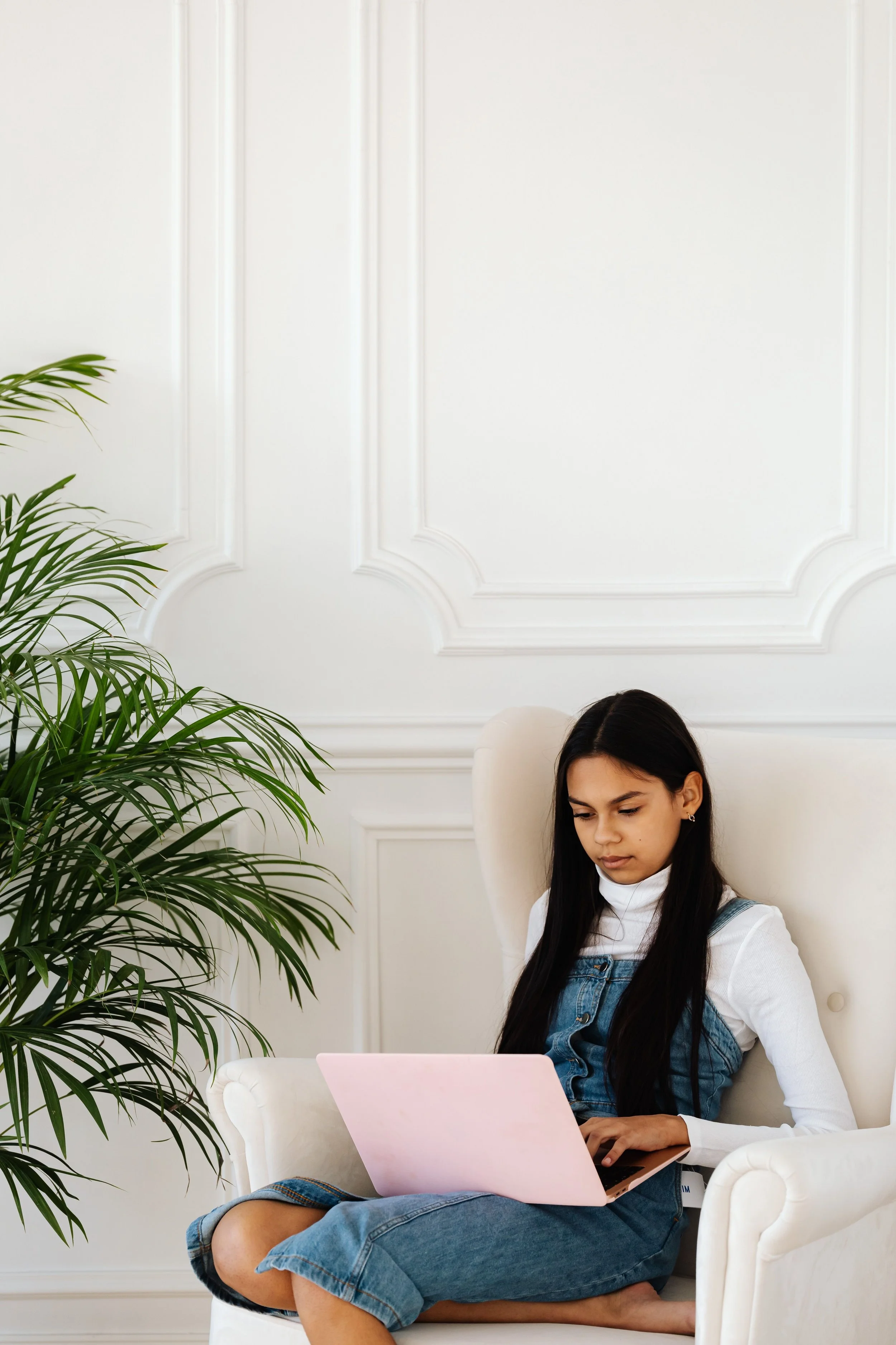 Young woman with long dark hair sitting cross-legged on a white armchair, using a pink laptop in a bright, elegant room with white paneled walls and a large green plant nearby.