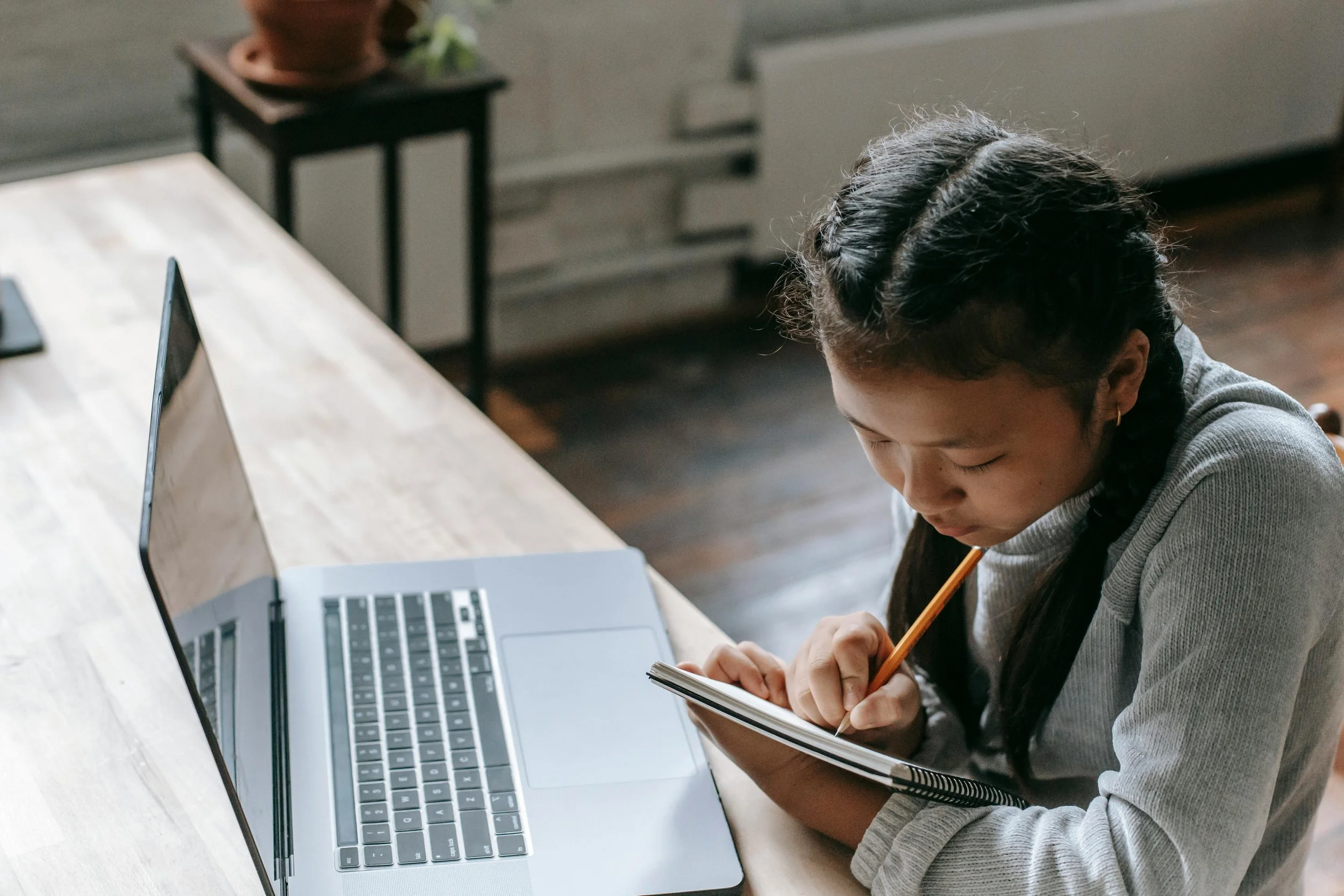 A young girl with braided hair, wearing a gray sweater, sitting at a wooden table with a laptop computer and sketchbook, writing with a pencil.