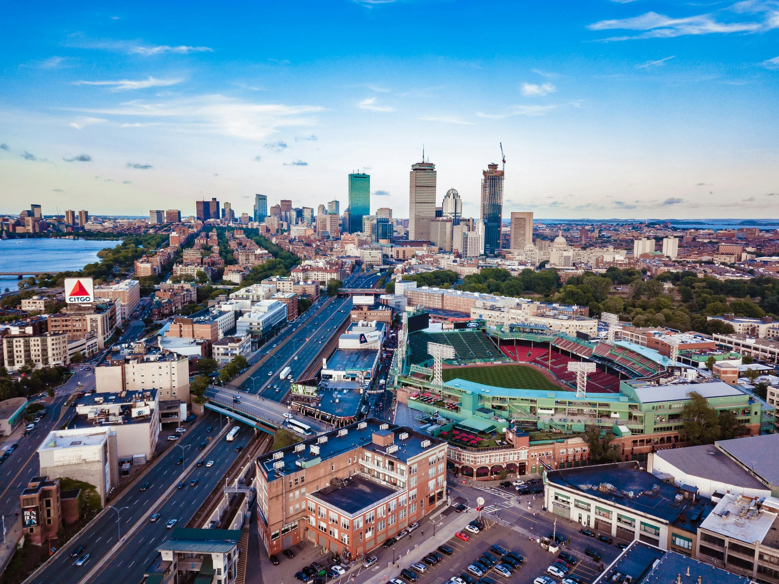 Aerial view of downtown Boston with skyscrapers, highway, and Fenway Park stadium with red seating.