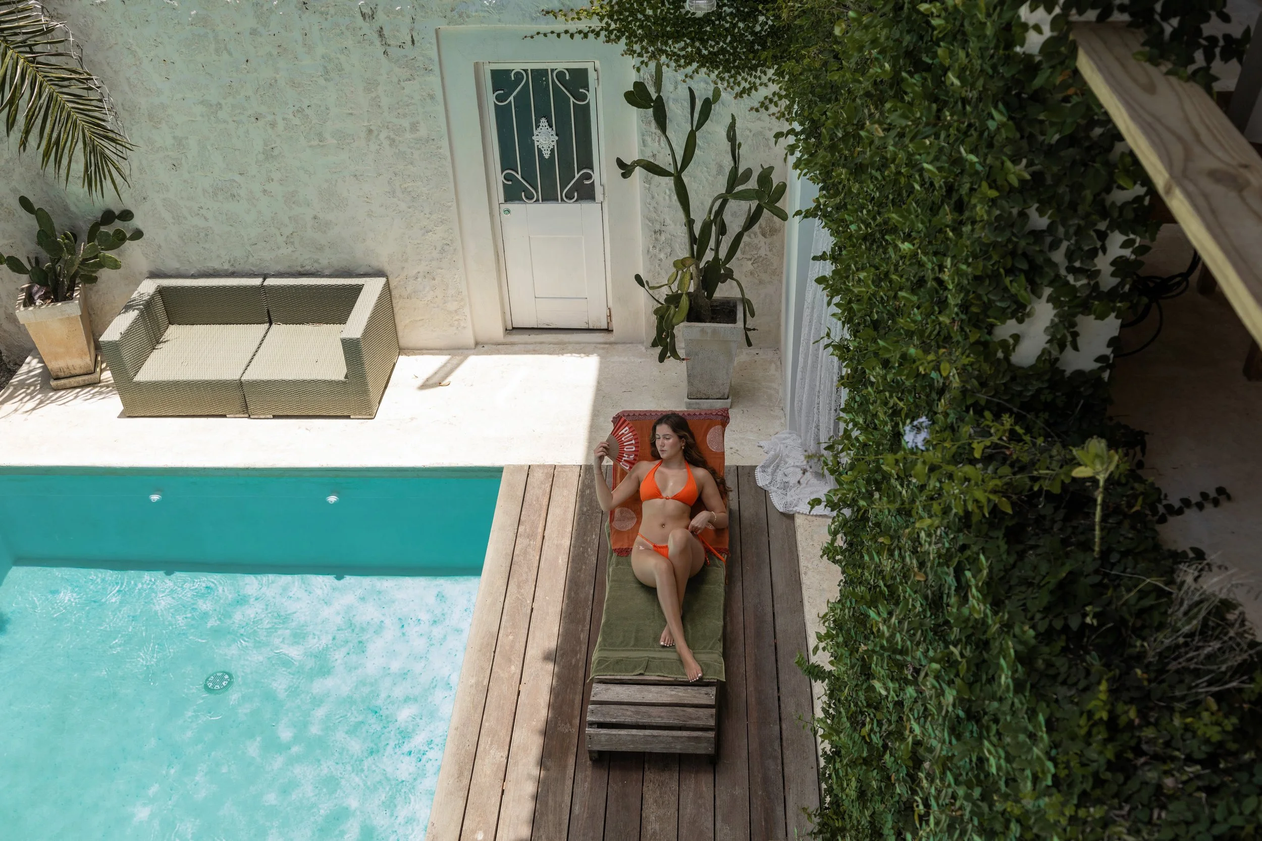 A woman in an orange bikini relaxing on a lounge chair beside a swimming pool, holding a fan, with greenery and a white door in the background.