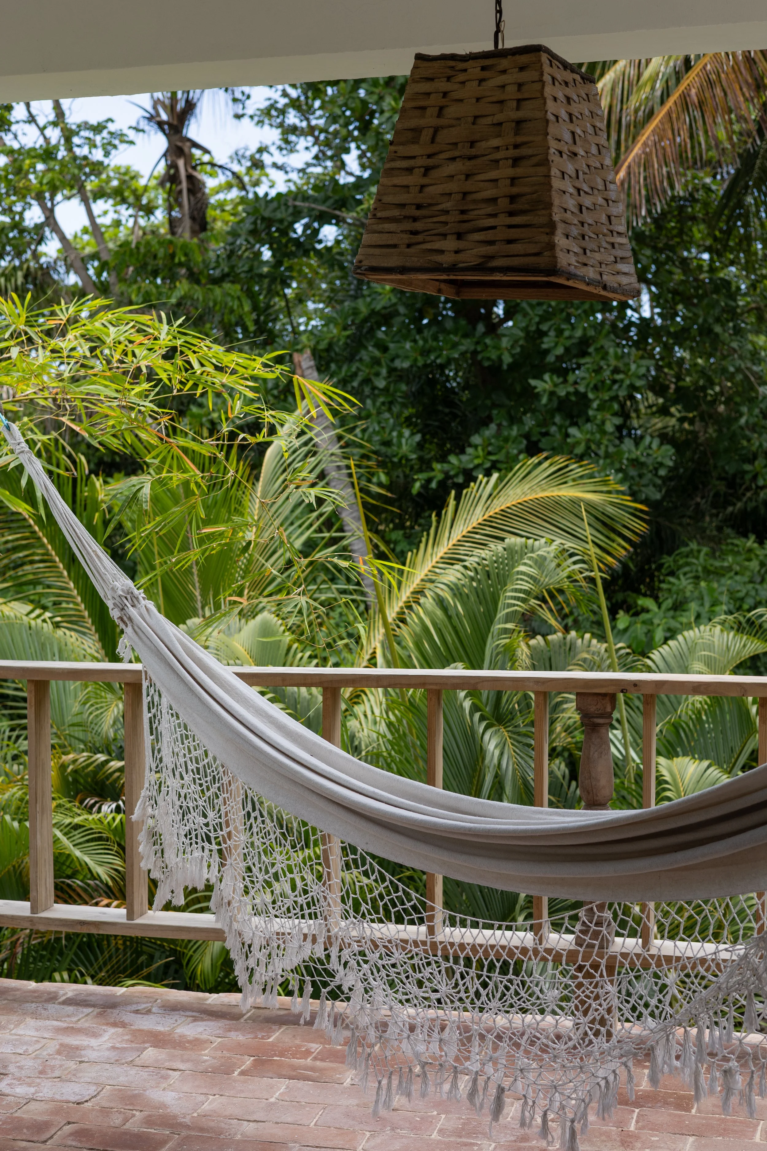A hanging wicker lamp on a porch with tropical plants in the background, a white hammock with fringe, wooden railings, and a brick floor.