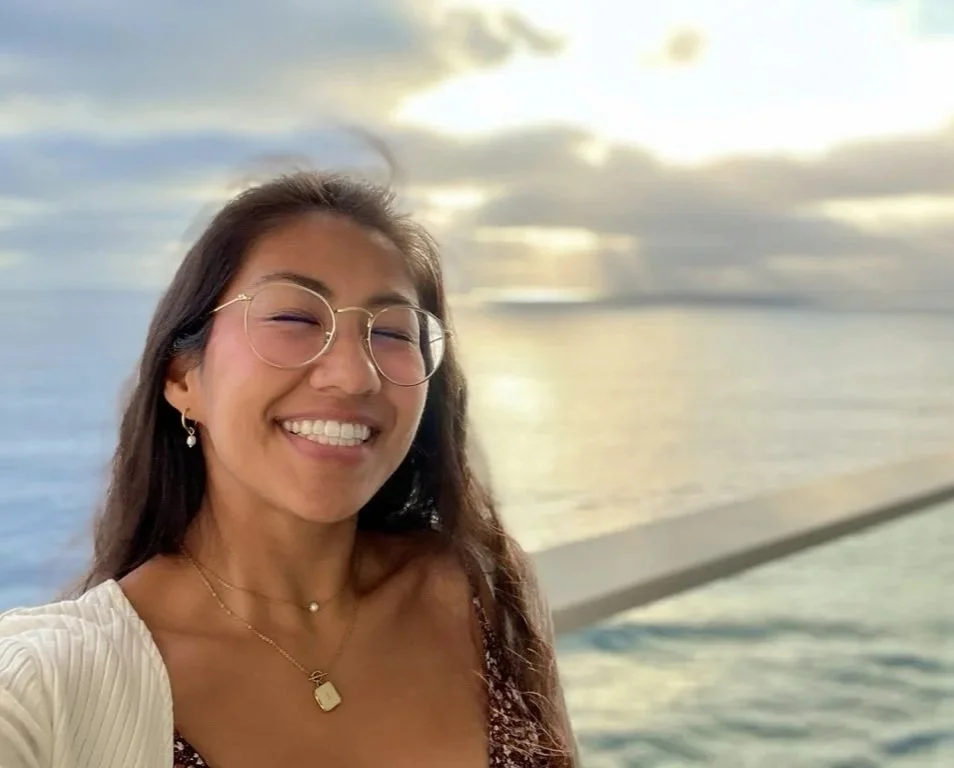 A young woman with glasses and earrings taking a selfie near a body of water during sunset.