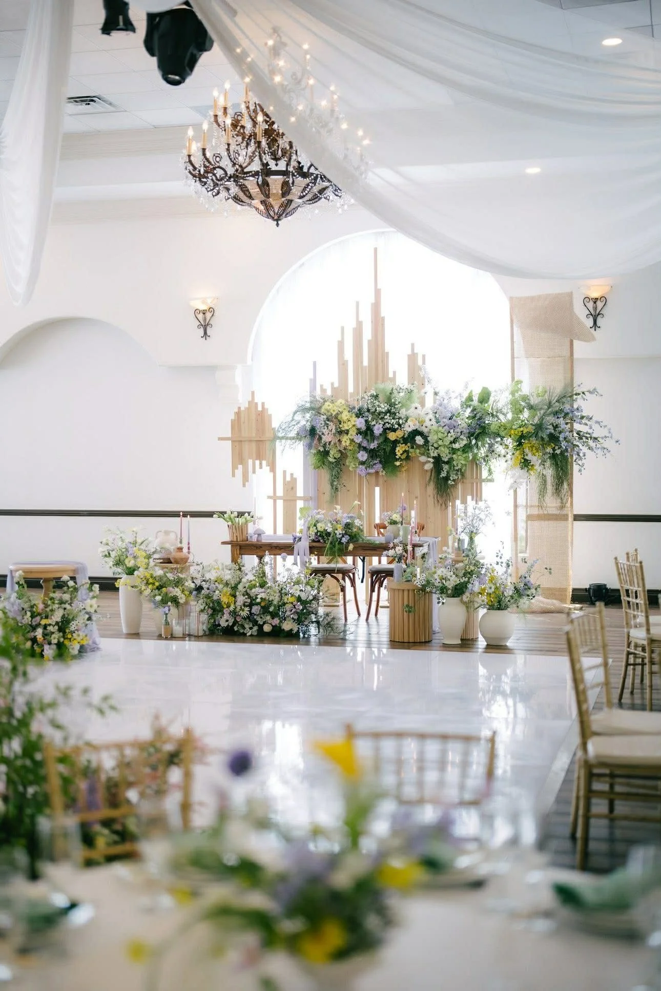 A wedding ceremony setup with floral decorations, a chandelier, and a wooden backdrop in a bright, white room.