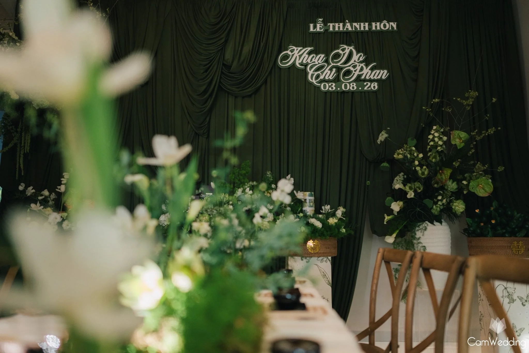 Indoor wedding ceremony setup with green curtains, floral arrangements, and a table with chairs in the foreground.