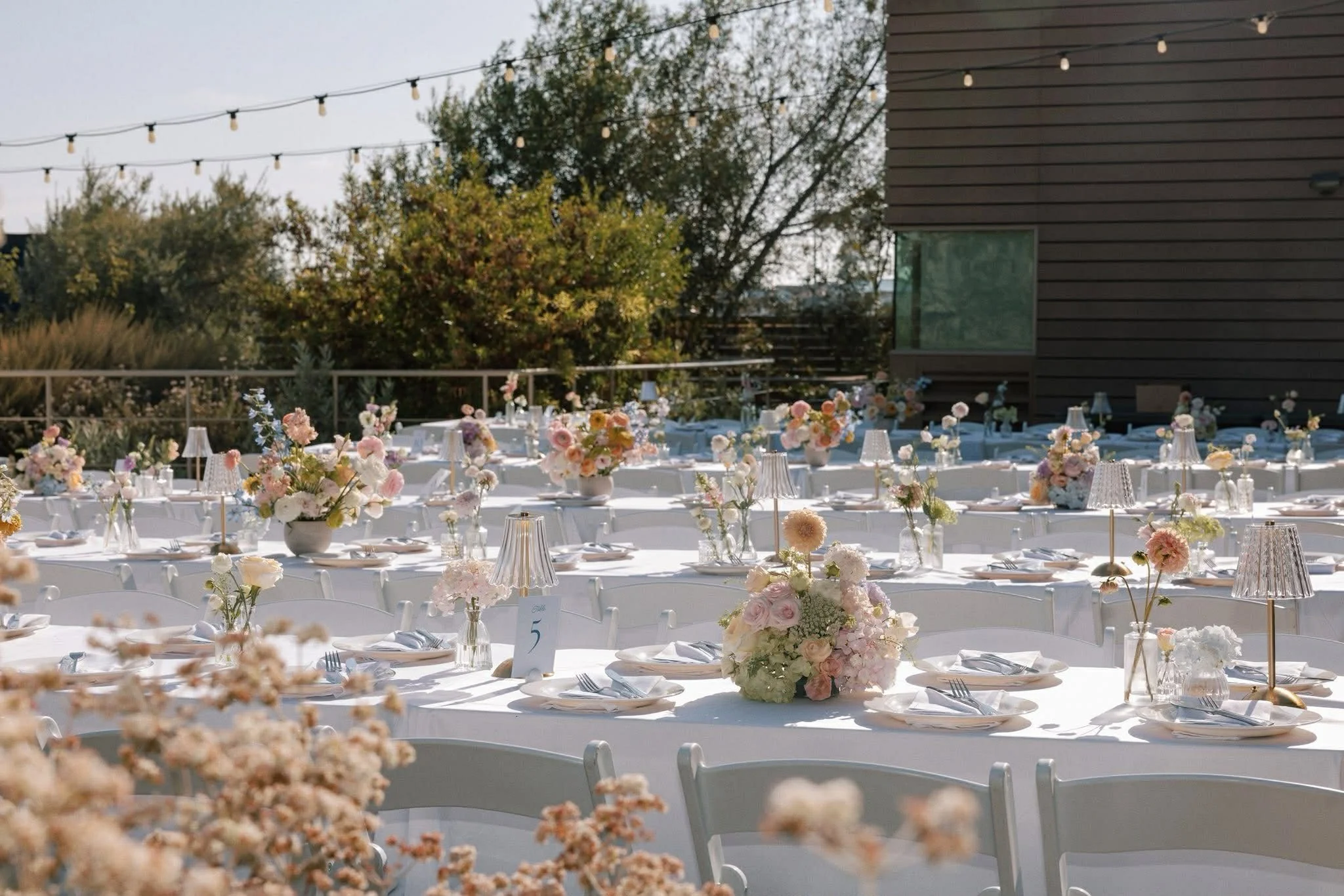 Outdoor wedding reception with round tables decorated with pastel-colored flower arrangements, white tablecloths, and elegant table settings during daytime.