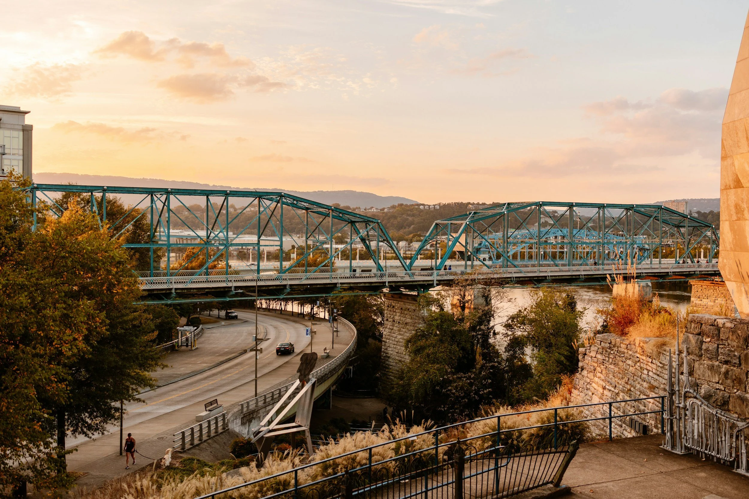 The Chattanooga Walnut Street Bridge, a steel-truss bridge over the Chattanooga river, at sunset with a hills in the background, cars on the bridge, trees on the riverbanks, and a road with a person walking a dog in the foreground.