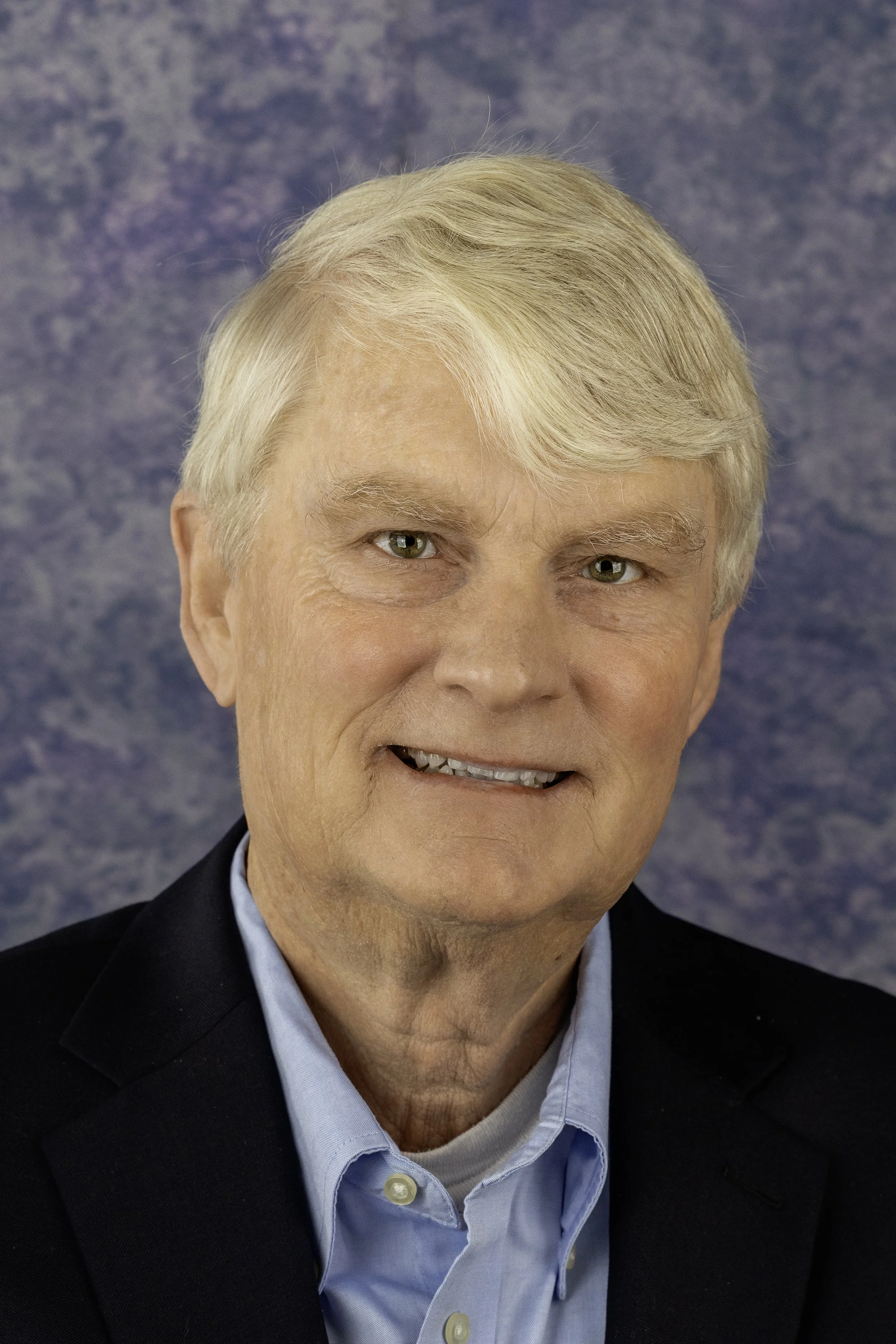 Close-up of a man with gray hair, wearing a blue shirt and a dark blazer, against a textured blue background.