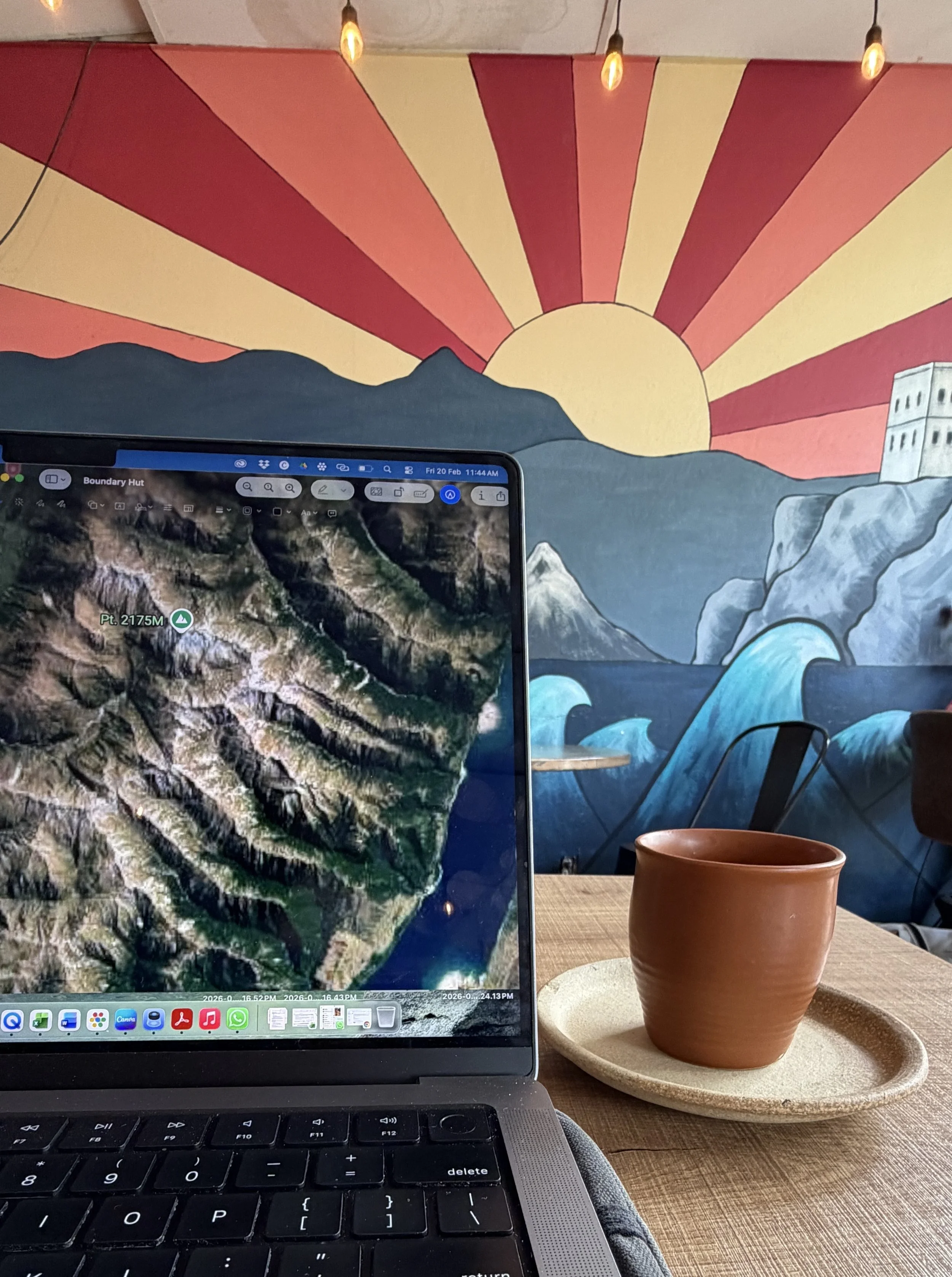 A laptop computer displaying a satellite map of a mountainous terrain, sitting on a wooden table next to a terracotta cup on a beige plate, with a colorful mural of a sunset over mountains, water, and waves in the background. Depicting work anywhere.
