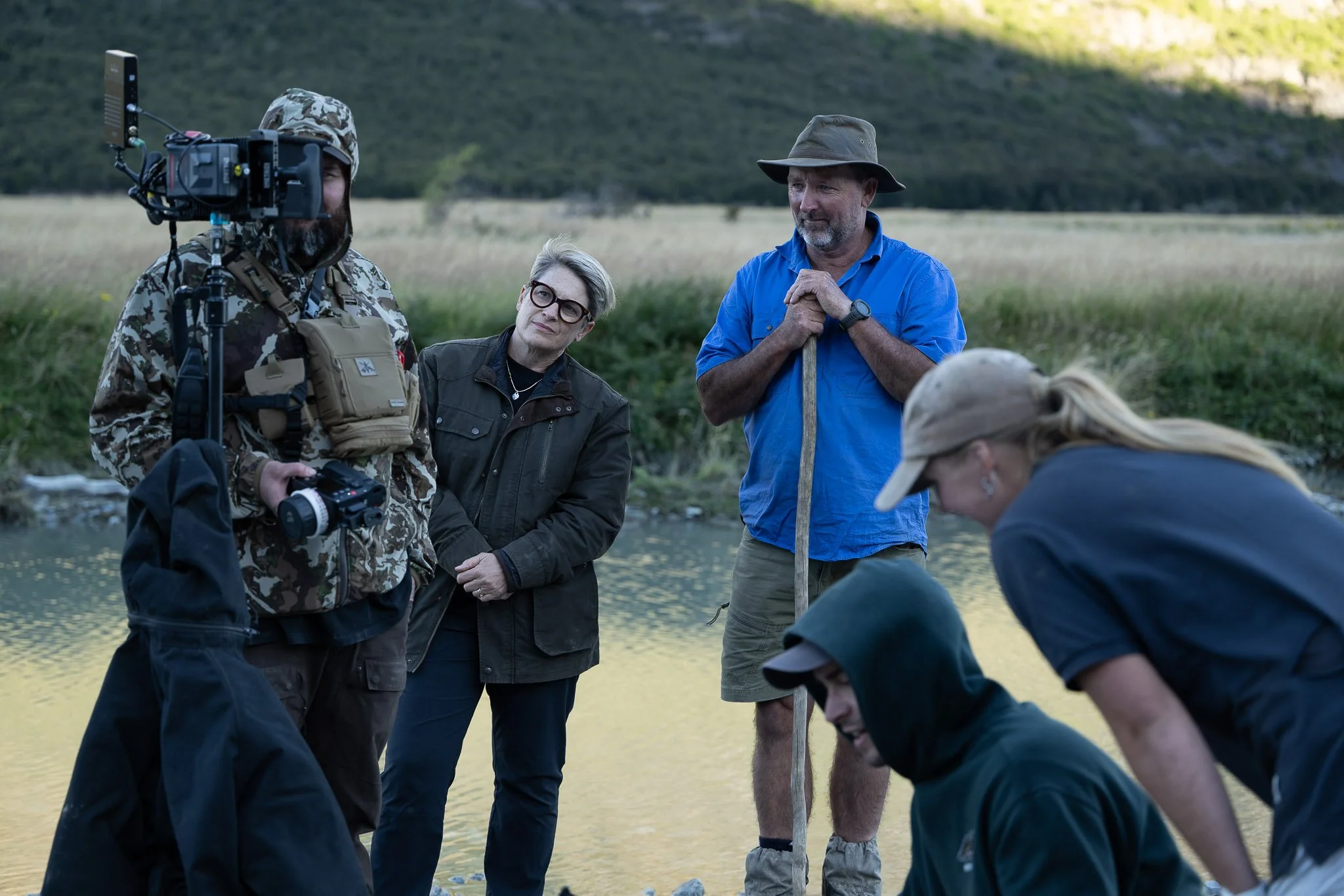 A group of people standing near a river outdoors, one person holds a camera on a stabilizer, others are engaged in conversation and looking at something on the ground, surrounded by grassy landscape.