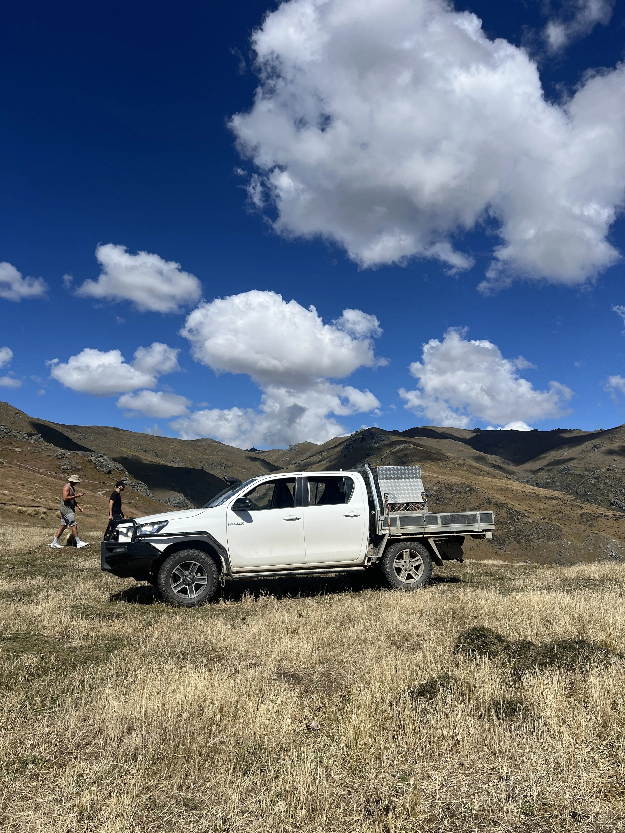 A white pickup truck parked on a grassy field in a mountainous area with two people walking nearby under a partly cloudy sky.