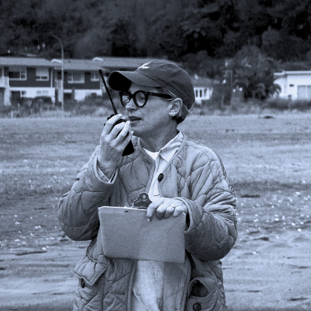 A woman wearing glasses, a cap, and a quilted jacket holding a clipboard and speaking into a walkie-talkie on set at the beach with crew, camera focus puller and director, with houses in the background.