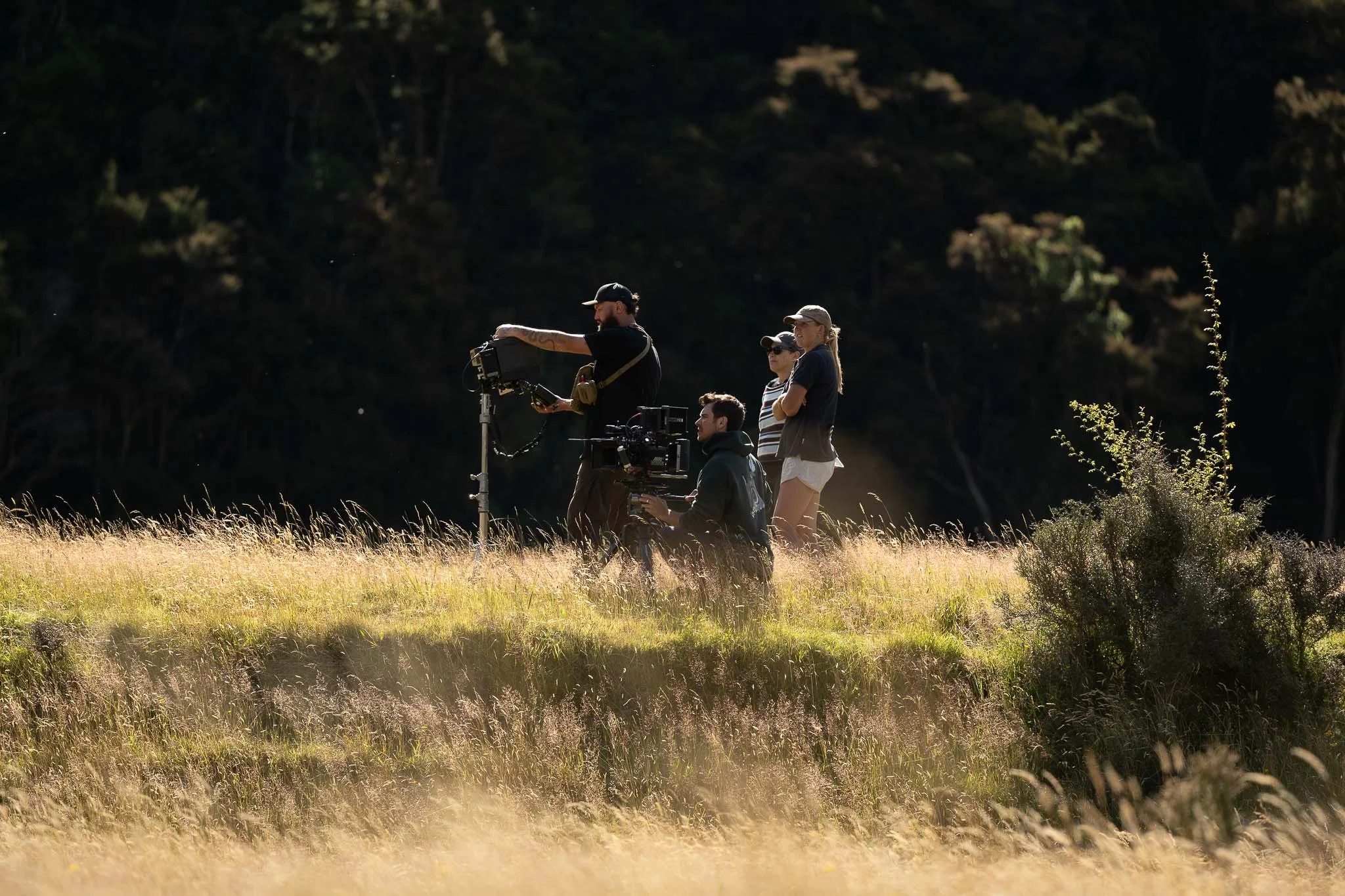 Film crew working outdoors in a field, with three people standing and one person operating a camera, as sunlight shines on the grass and trees in the background. Small crews, travelling anywhere you need to capture photos or footage.