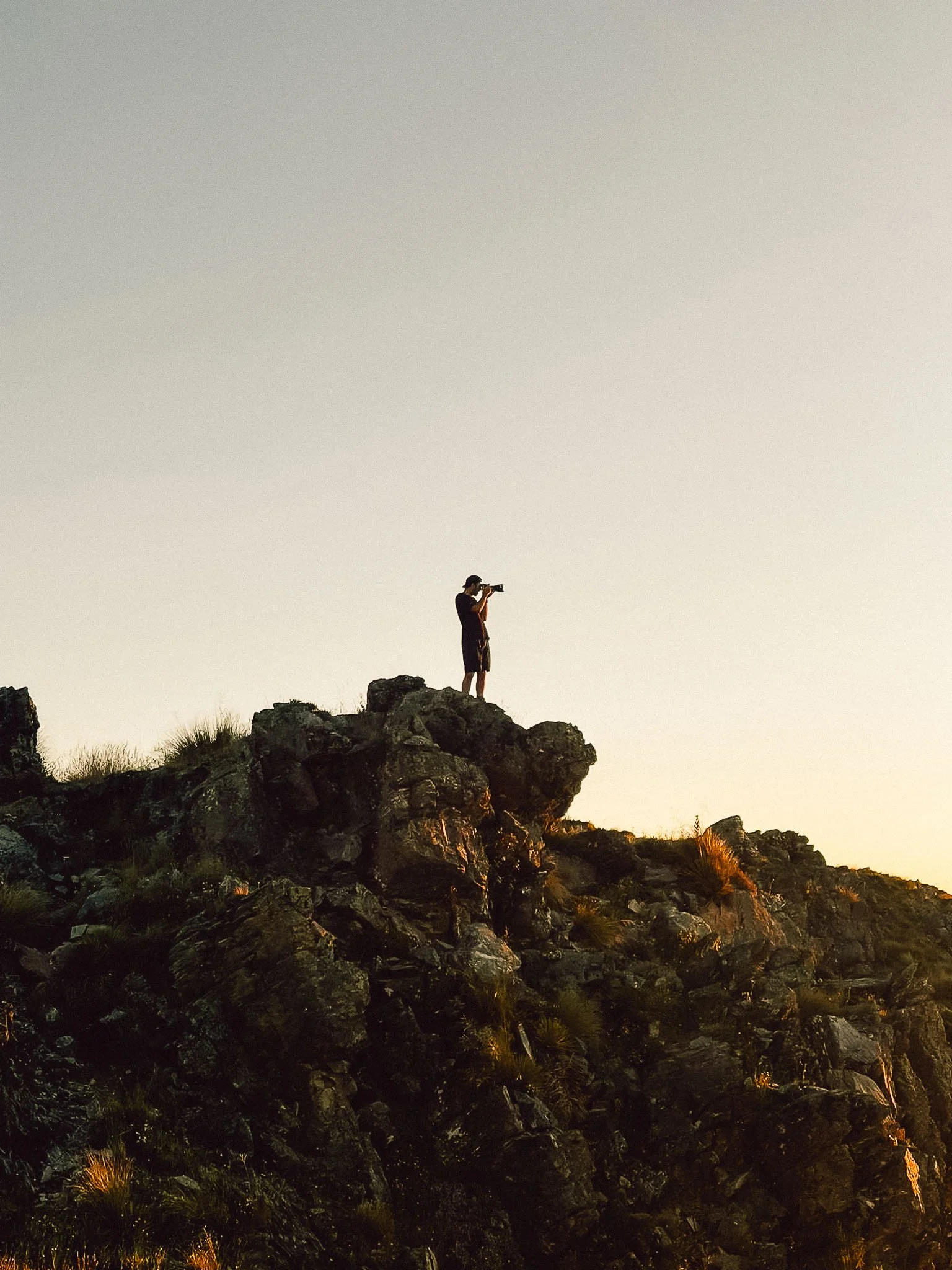Photographer standing on top of a rocky hill or mountain in South Island, New Zealand high country, during sunset or sunrise, taking a photograph with a camera. Canon camera expert. Sony photography expert.