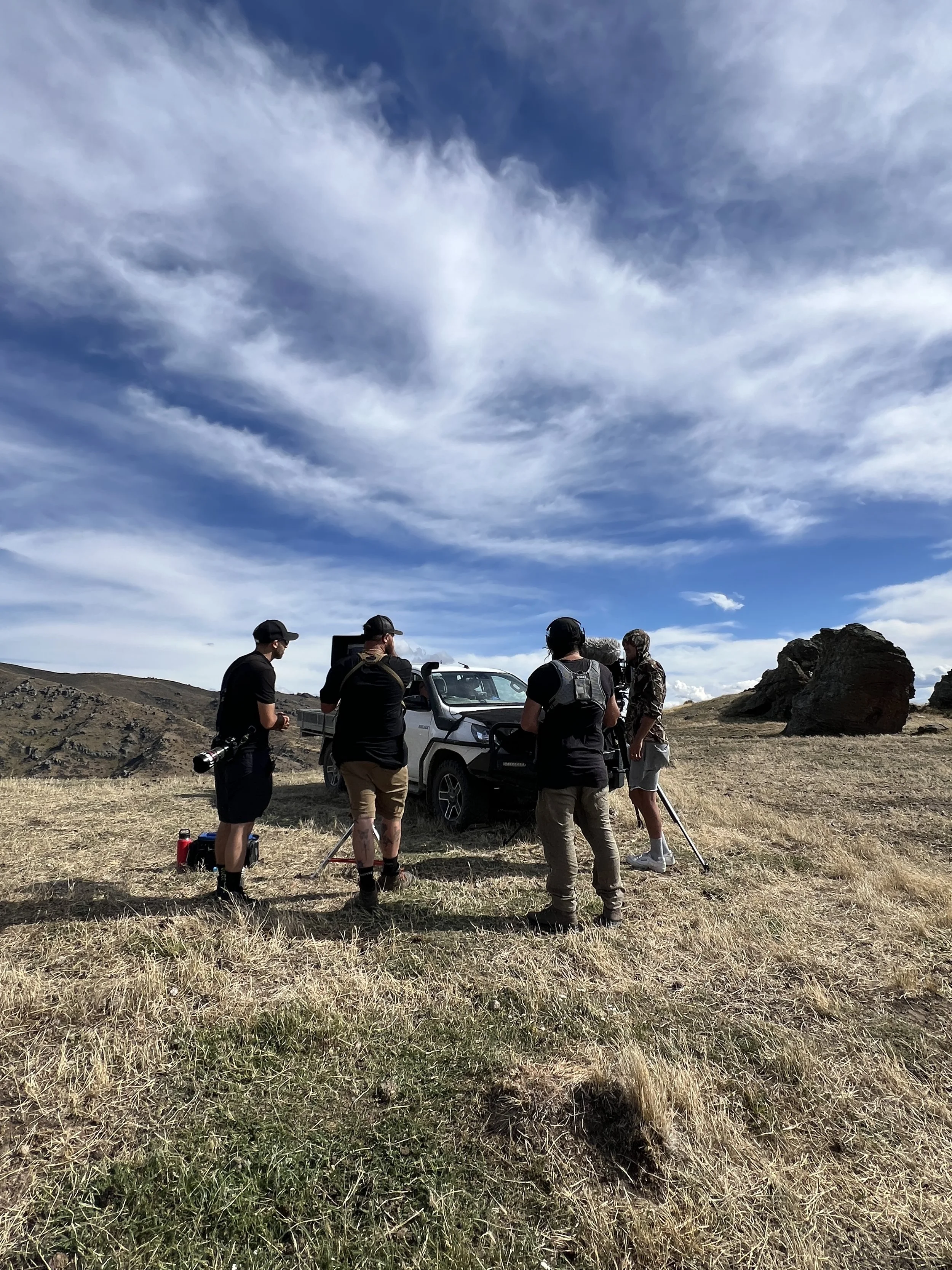 Group of five people standing beside a vehicle in a grassy, hilly landscape with large rocks and a partly cloudy sky.
