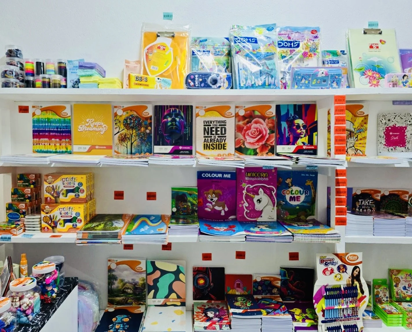 A retail store shelf filled with colorful notebooks, art supplies, and stationery items, including drawing books, coloring books, and craft kits, with some items featuring animated characters and vibrant designs.