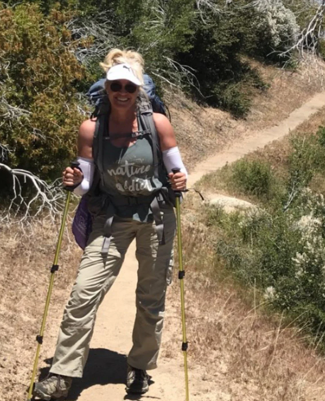 Person hiking on a trail with trekking poles, wearing a backpack and visor.