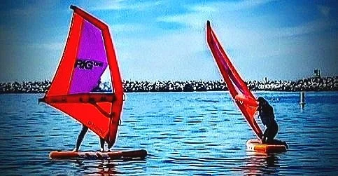 Two people in wetsuits near an inflatable orange watercraft, one holding a standup paddle board with a sail-on the water.