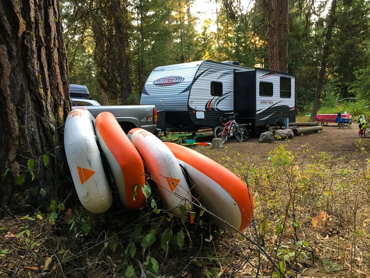 A forest campsite with a travel trailer, bicycles, and paddle board rentals by Play Fair Paddleboarding, surrounded by trees and outdoor gear.