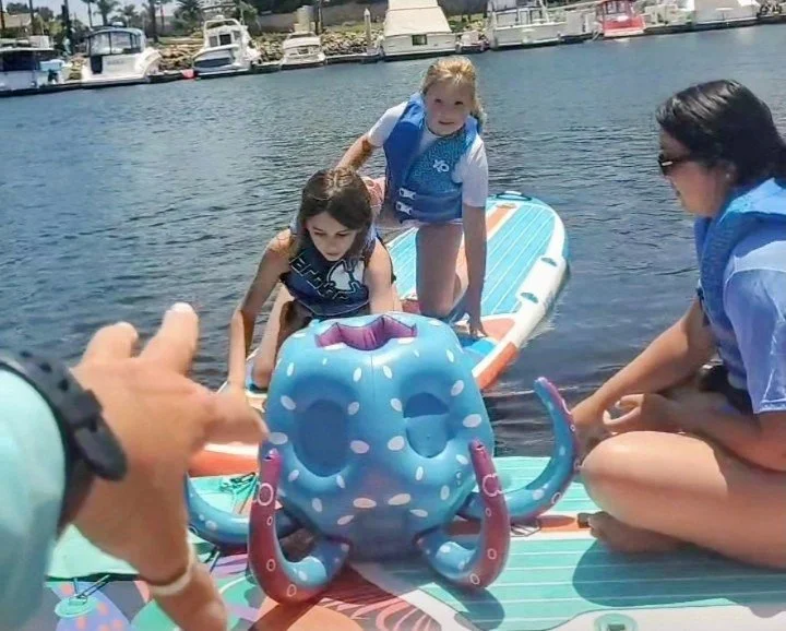 YMCA day camp session with instructor Julie Jenkins, on a dock with a paddleboard and a blue octopus-shaped pool float, with boats and a marina in the background.