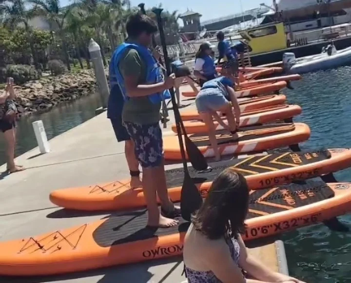 People preparing stand-up paddleboards at a marina dock, with some standing and others bending over to adjust or check the boards.  Fleet of paddle board rentals from Play Fair Paddleboarding- services - Lake Hartwell