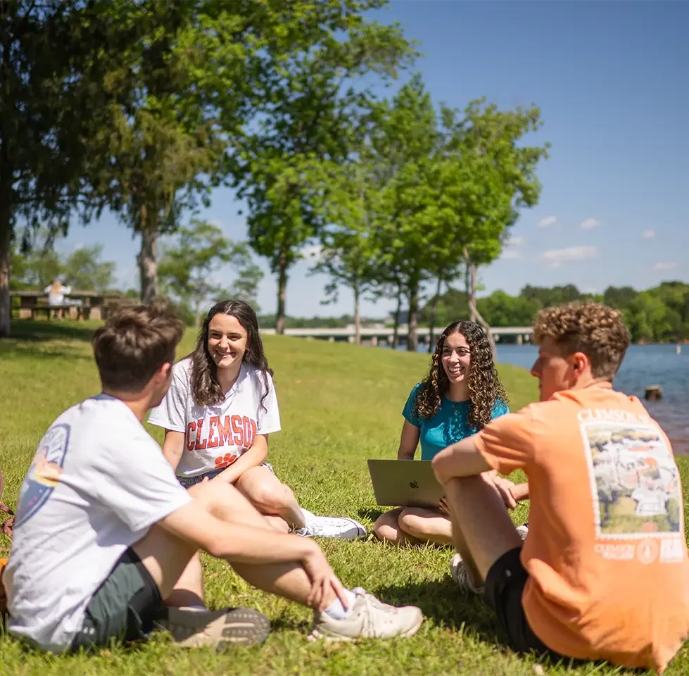 Four clemson students sitting on grass by lake hartwell, smiling and talking on a sunny day, with trees and a bridge in the background. Paddle Board rentals - Play Fair Paddle Boarding - lake Hartwell