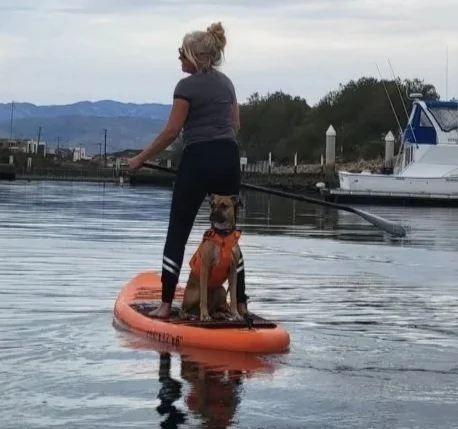 Owner of Play Fair Paddleboarding with her dog on an orange board in a calm body of water, with boats and mountains in the background.