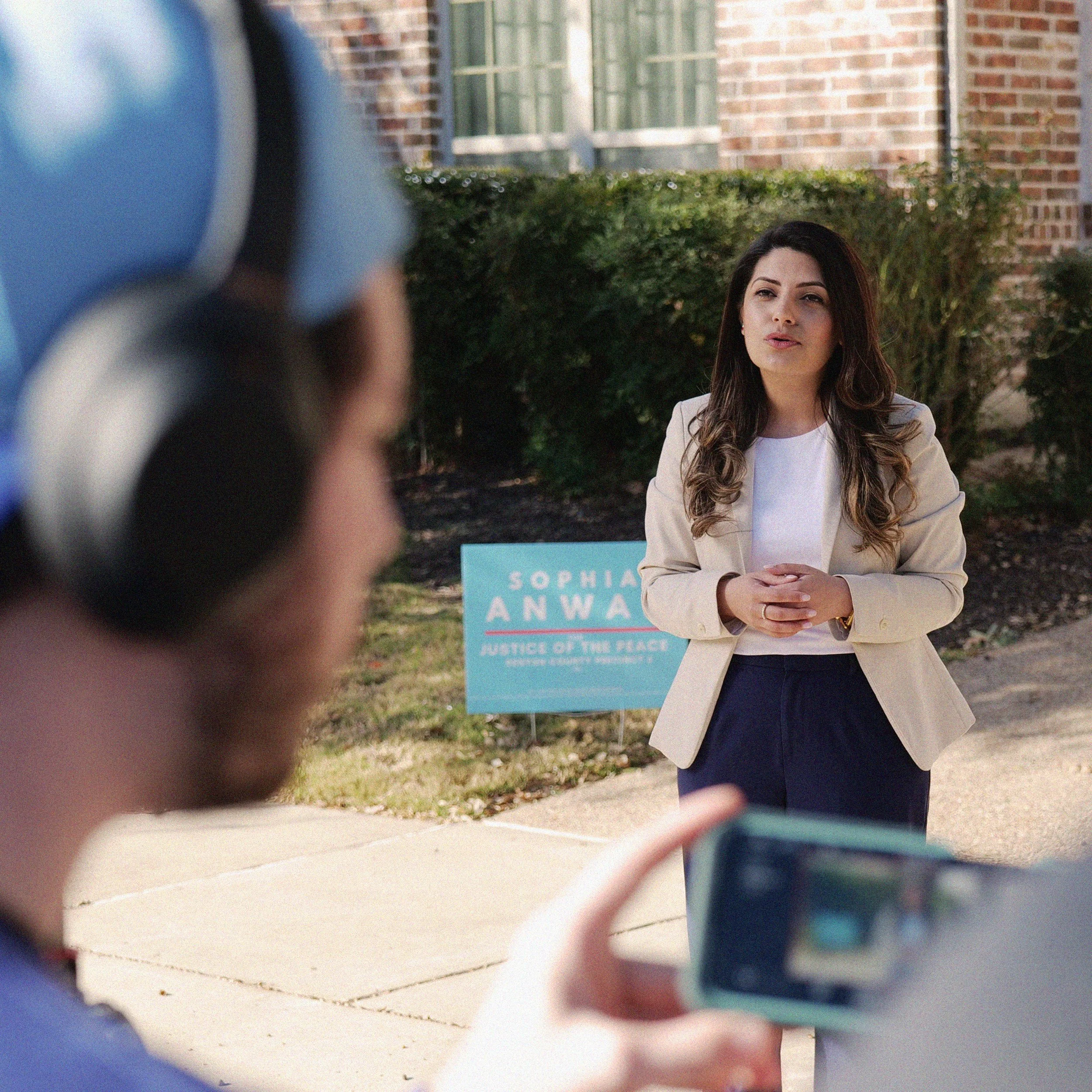 A woman with long dark hair and a beige blazer sitting outdoors in front of a bush and brick building, during a daytime interview or conversation, viewed through a blurred person's shoulder and hair in the foreground.