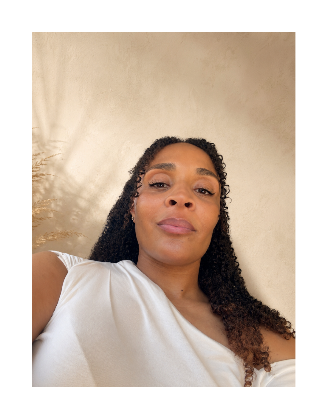 Close-up selfie of a woman with curly hair, wearing a white top, looking at the camera against a neutral wall background.