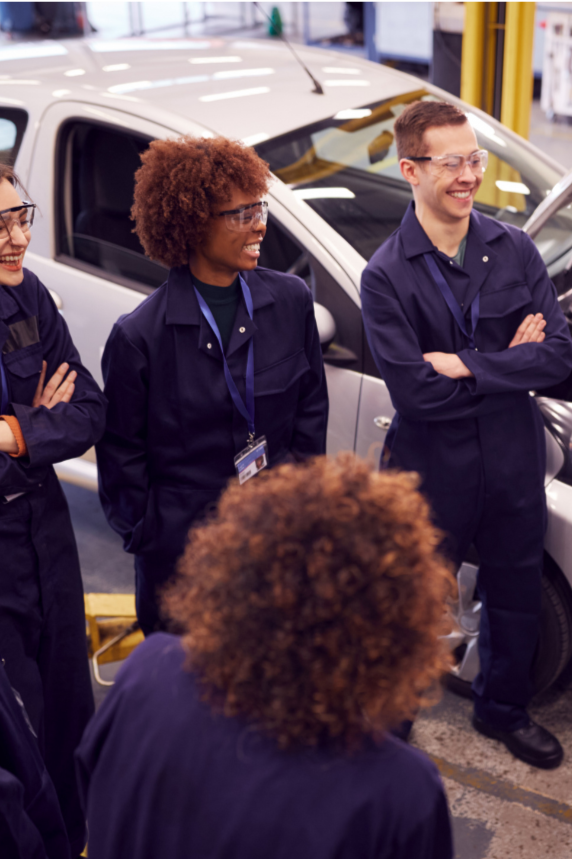Group of employees in safety gear standing near a car in a parking lot, smiling and talking.