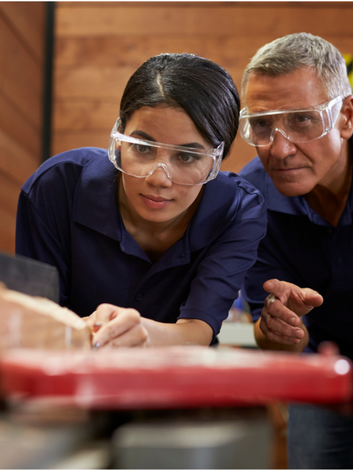Two people, one woman and one man, wearing safety goggles and navy blue shirts, are focused on an experiment or project in a woodworking or laboratory setting, with wooden walls in the background.