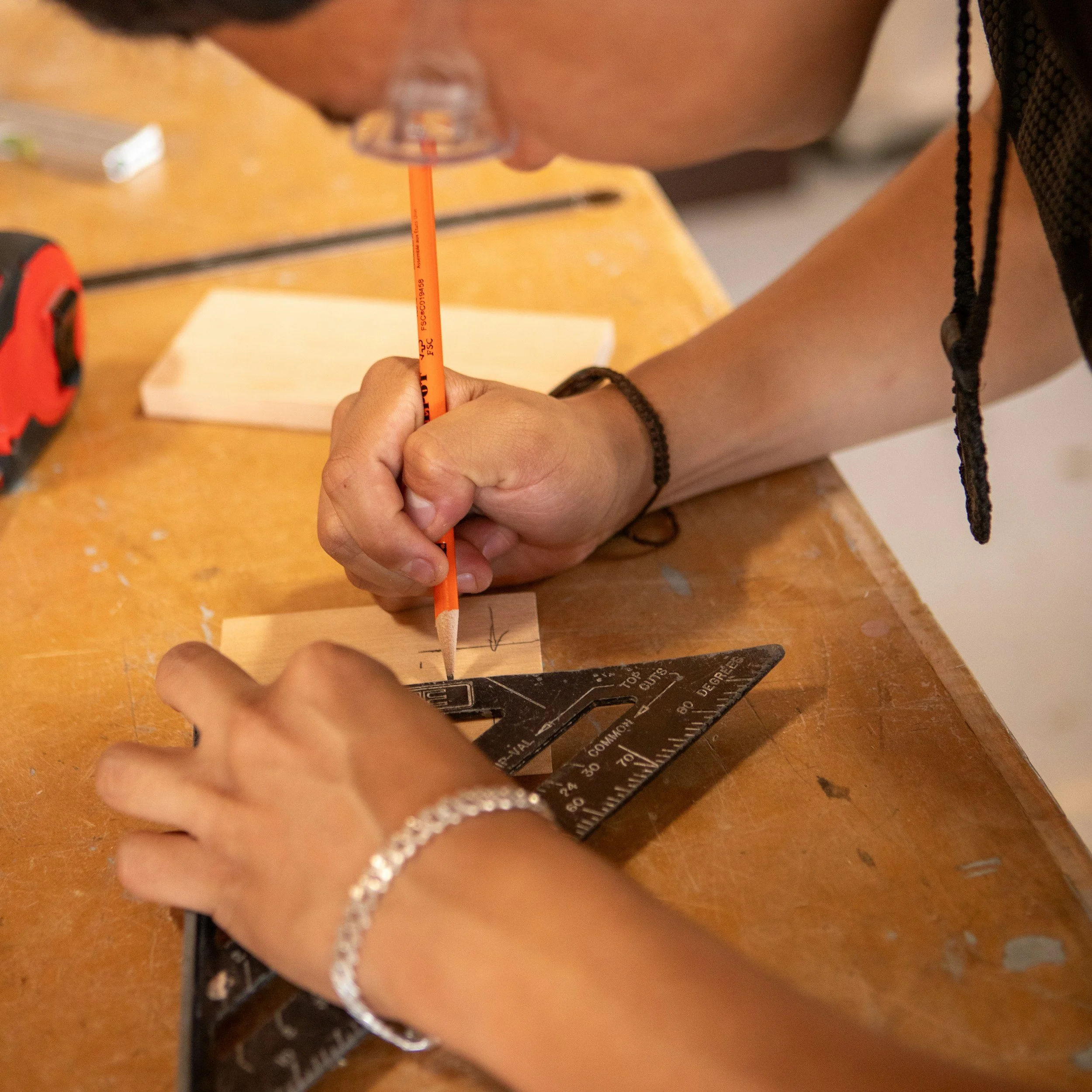 Close-up of a person’s hand holding a pencil and drawing on a piece of paper, with a black protractor and measuring tools on a wooden desk.