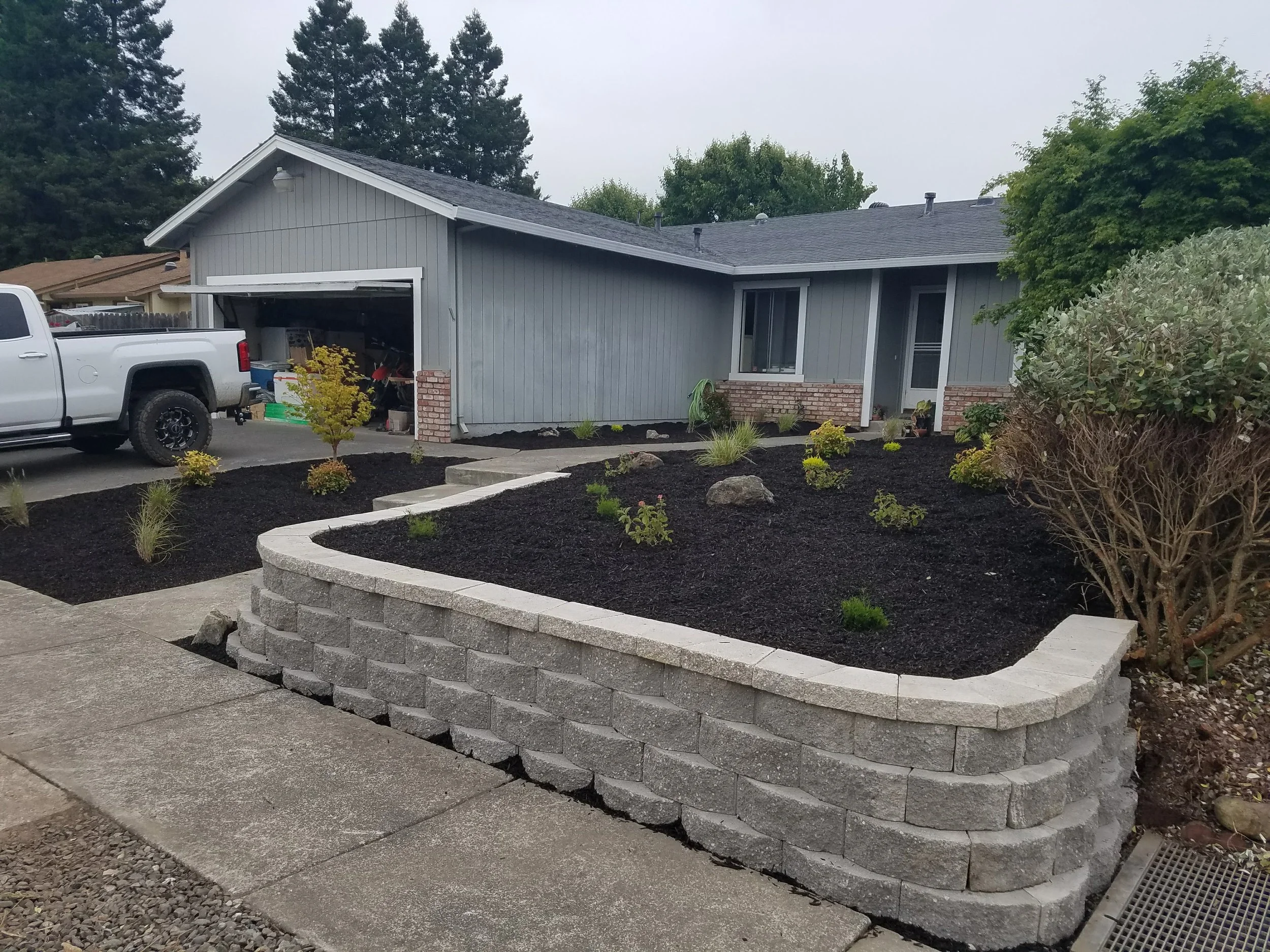 A house with gray siding and a dark roof, with a partially open garage and a white pickup truck parked in the driveway. The front yard has small shrubs and a curved brick retaining wall, with a patch of freshly laid dark mulch and some plants. There are tall trees in the background and an overcast sky.